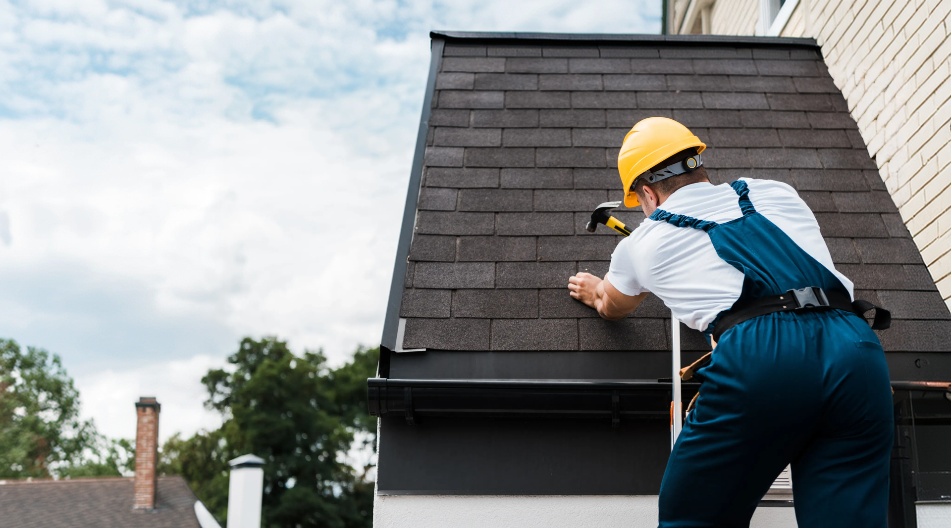 Roofer in a yellow hard hat hammering asphalt shingles into place on a steep roof