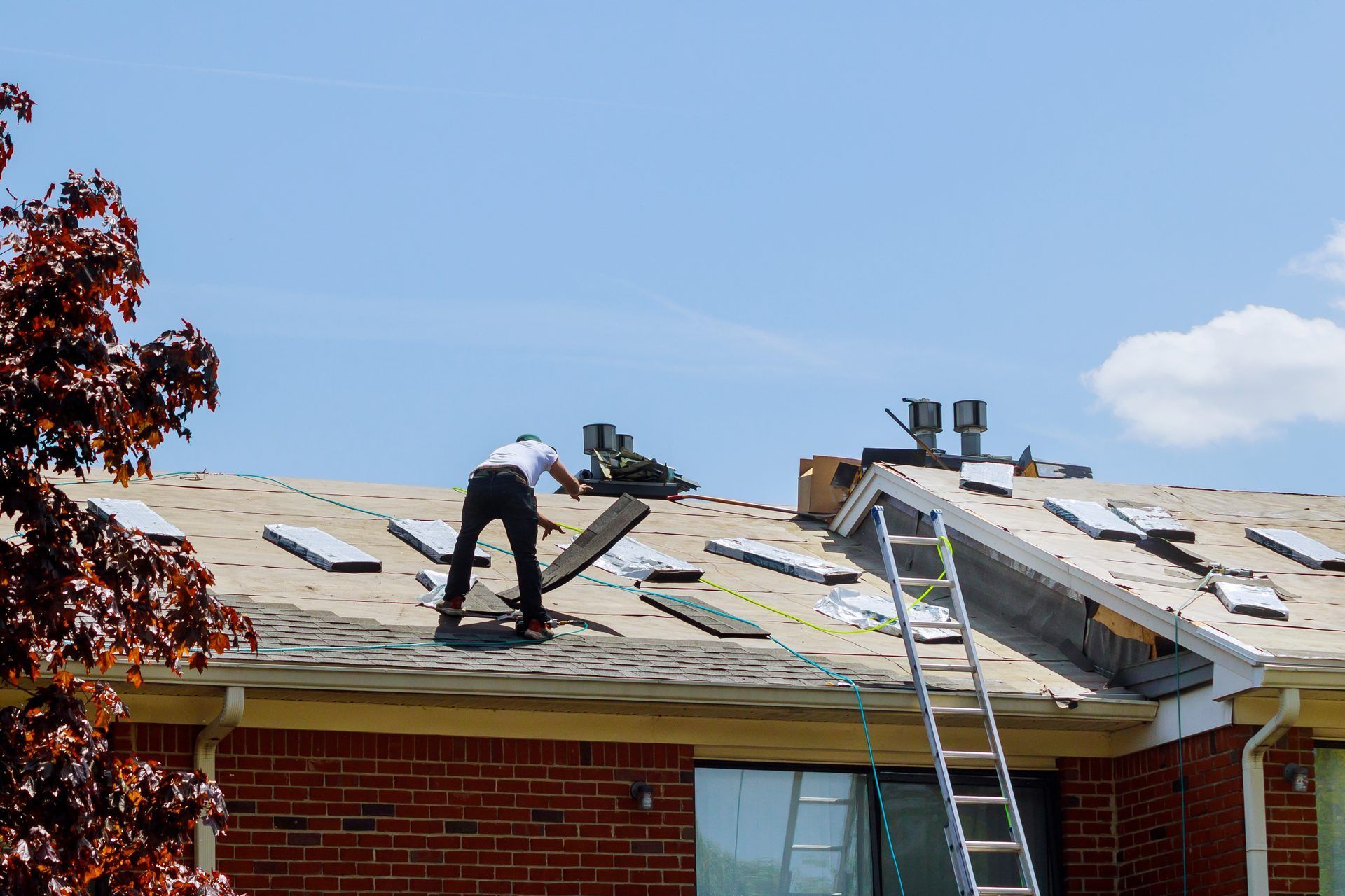 Roofing worker stripping old shingles from a brick house with a ladder propped against the eave