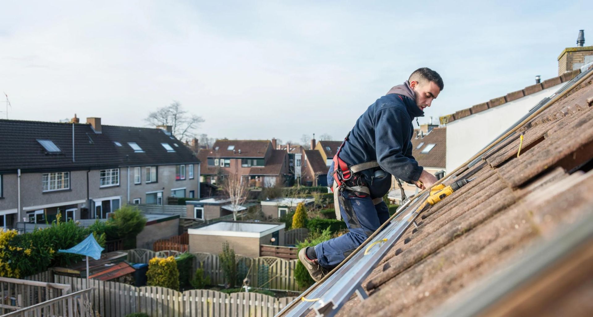 Roofer in a safety harness fastening mounting rails to a tiled roof with a power drill