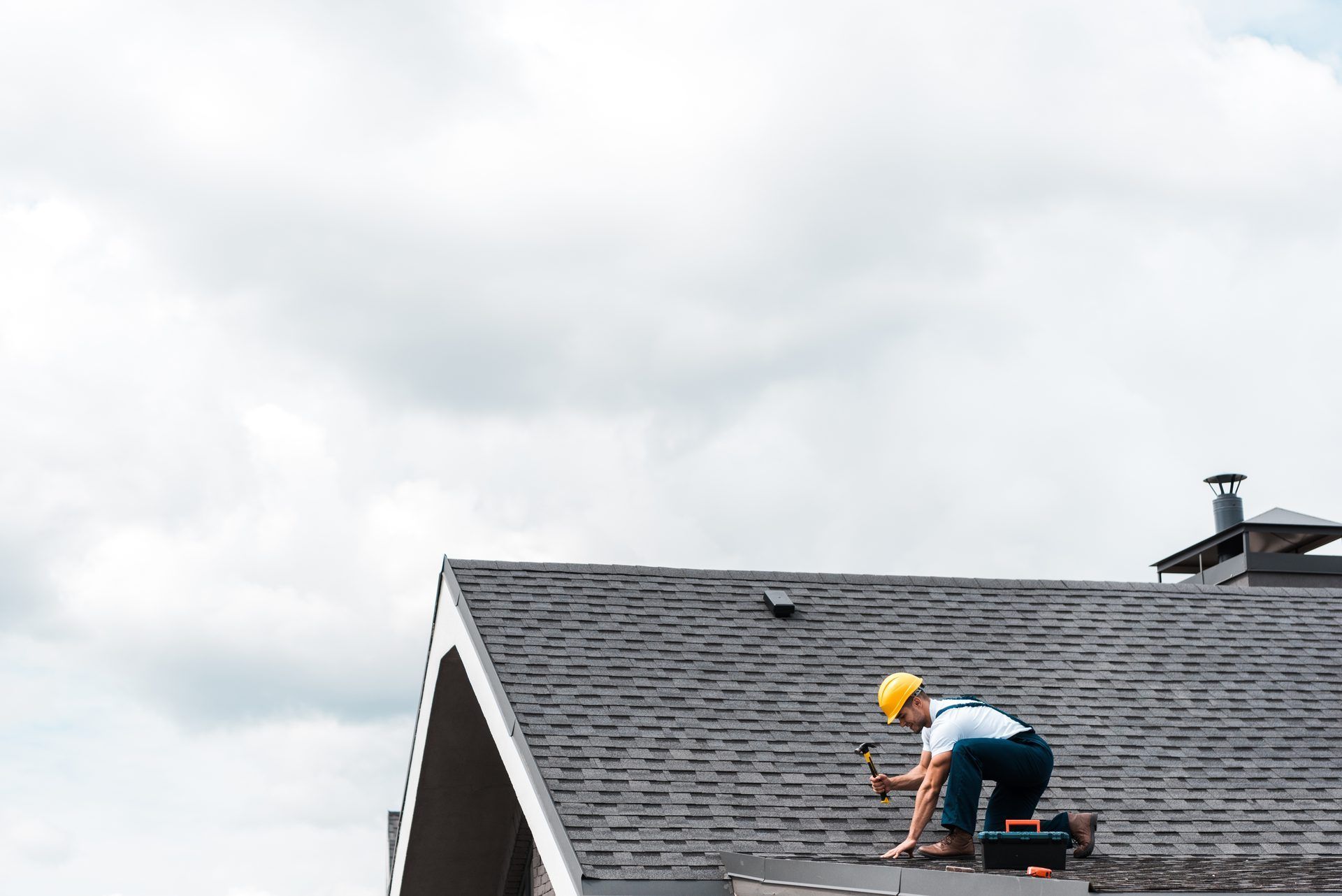 Roofer in a yellow hard hat working with a hammer near the ridge of an asphalt shingle roof