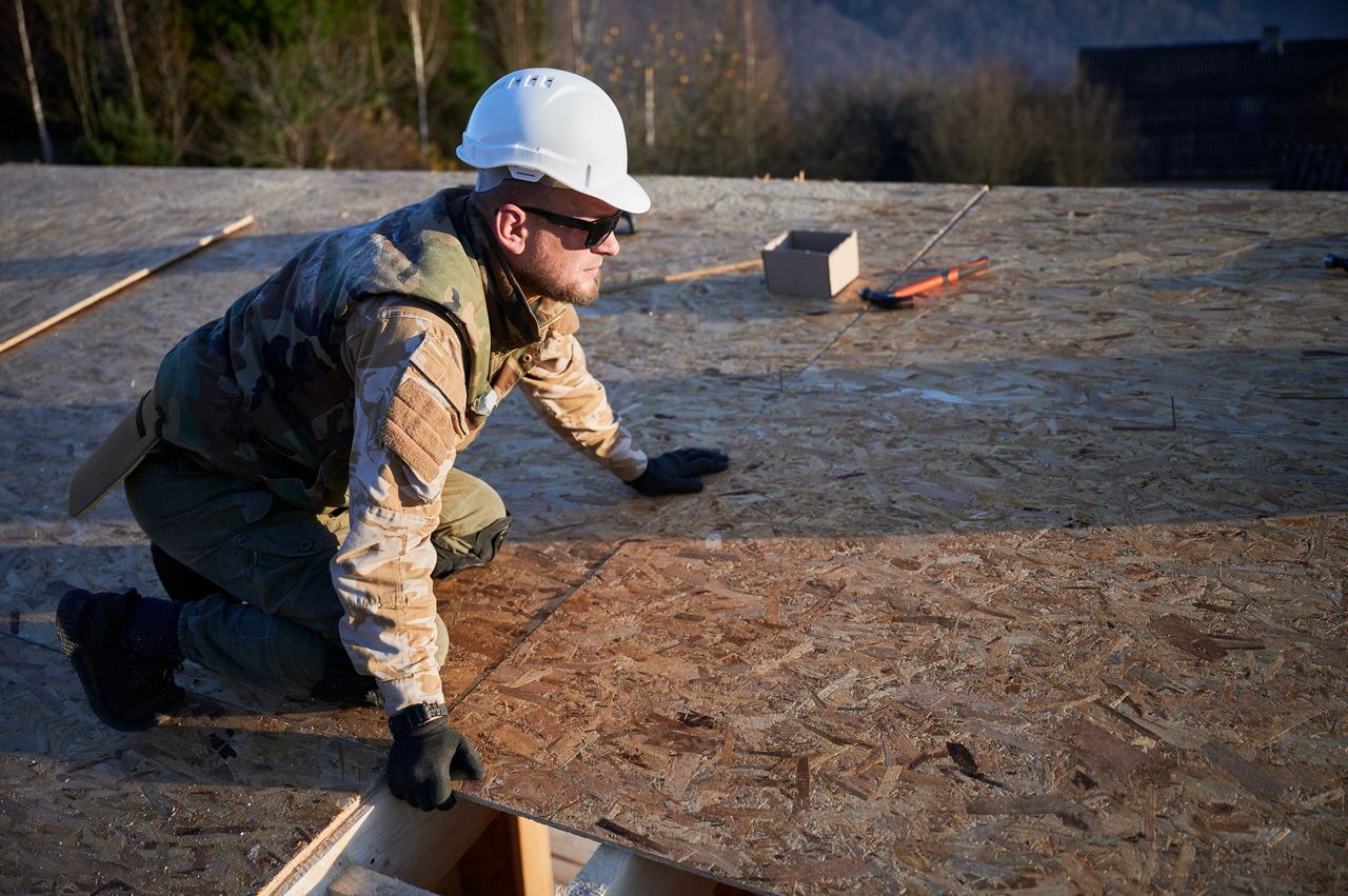 Roofer in a white hard hat kneeling on OSB roof sheathing during a jobsite evaluation