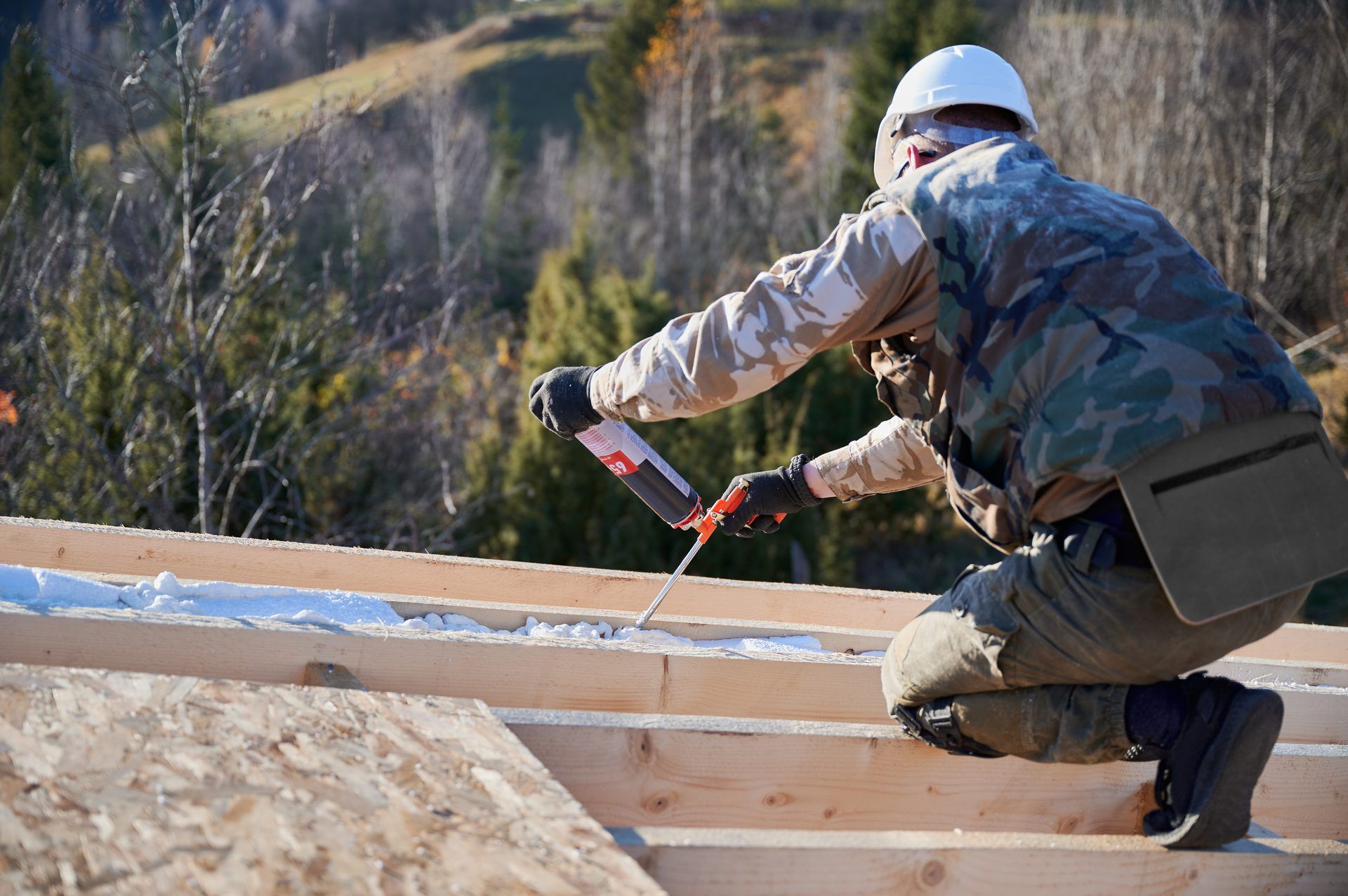 Roofer in a hard hat kneeling on a new timber roof frame, fastening boards with a nail gun