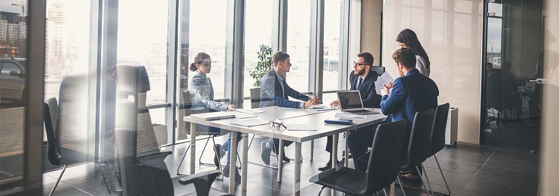 People seated around a glass conference table in a bright office meeting room, discussing and working.