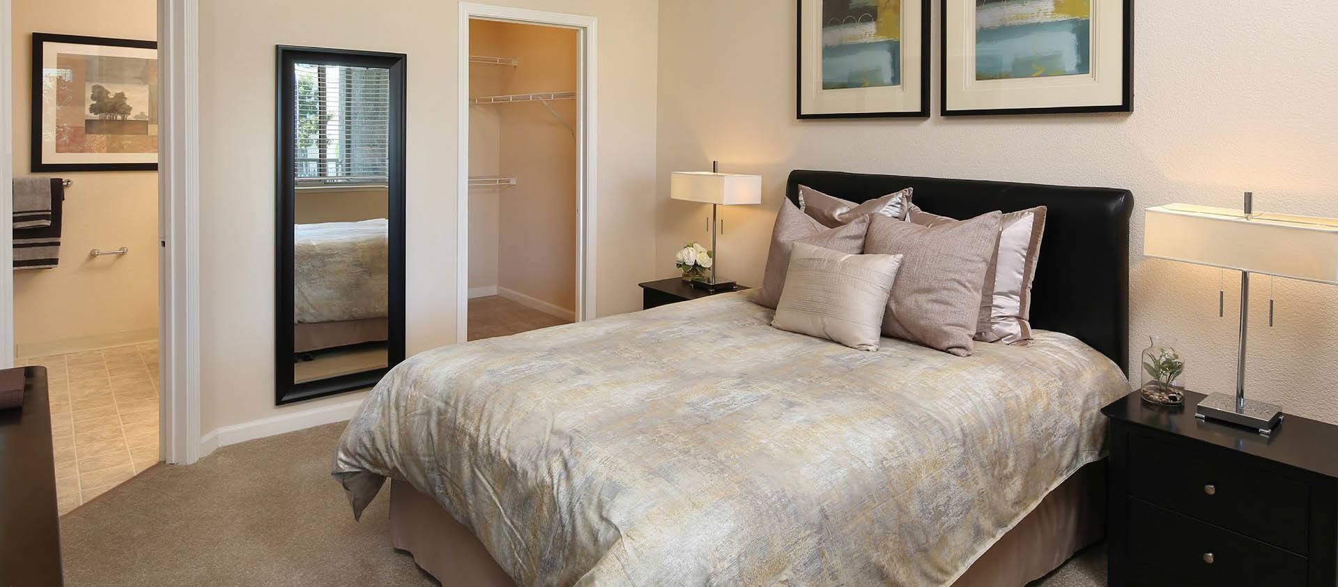 A bedroom with a bed, nightstands, a mirror, and an open closet. The walls are neutral colored at Iron Point at Prairie Oaks apartments in Folsom, CA.