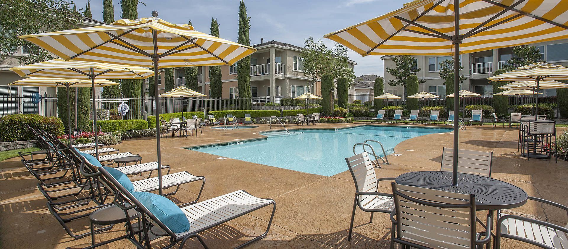 Poolside lounge area with striped umbrellas, chairs, tables, and a large building in the background at Iron Point at Prairie Oaks apartments in Folsom, CA.