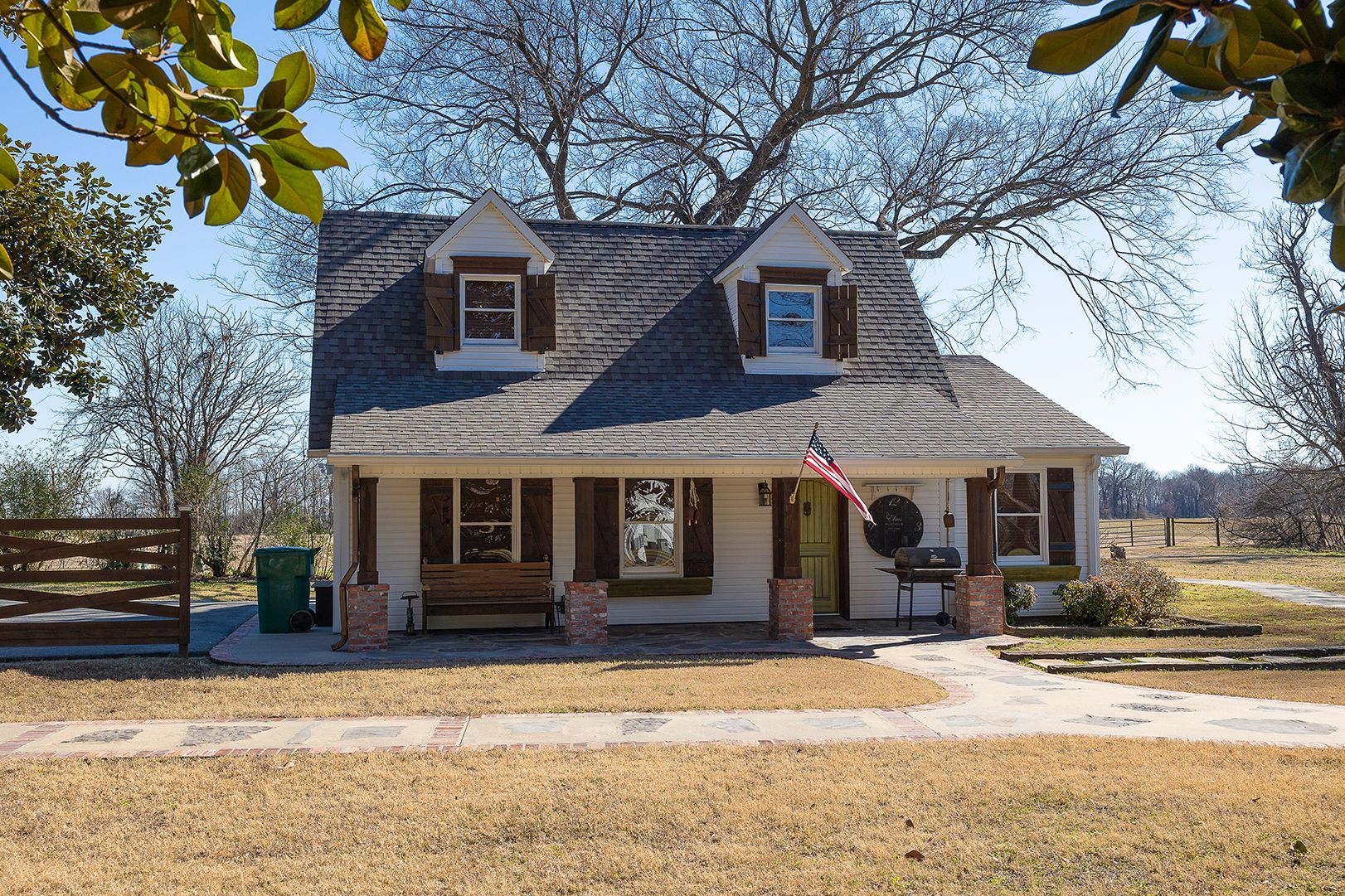 A white cottage with a grey shingled roof, two dormer windows, a front porch, and an American flag in a rural, grassy yard.