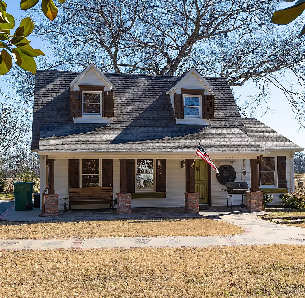 A white, two-story house with a shingled roof, two dormer windows, a front porch, and an American flag.