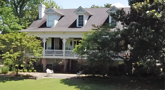 A two-story white house with a wraparound porch, three dormer windows, and a green lawn surrounded by trees.