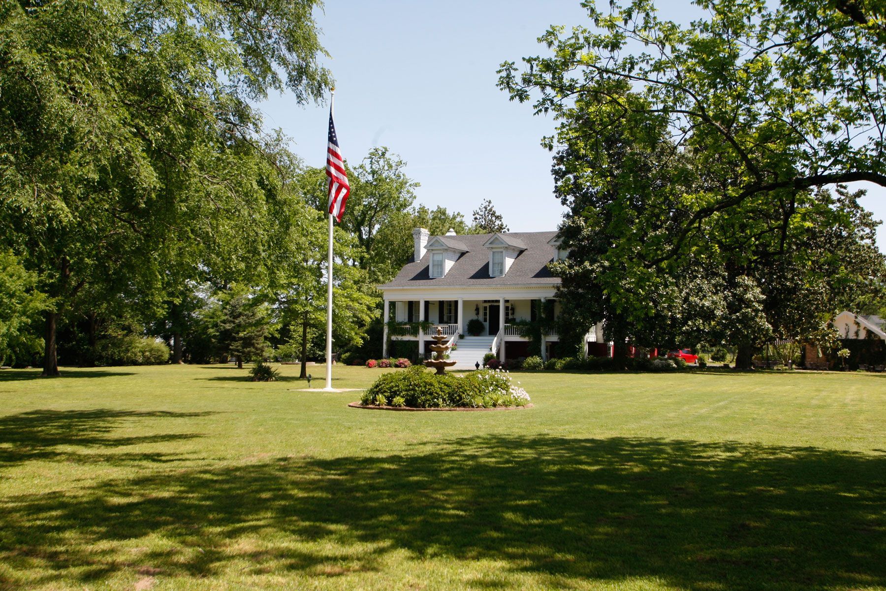 A white, two-story colonial-style house sits behind a large grassy lawn with an American flag flying on a pole.