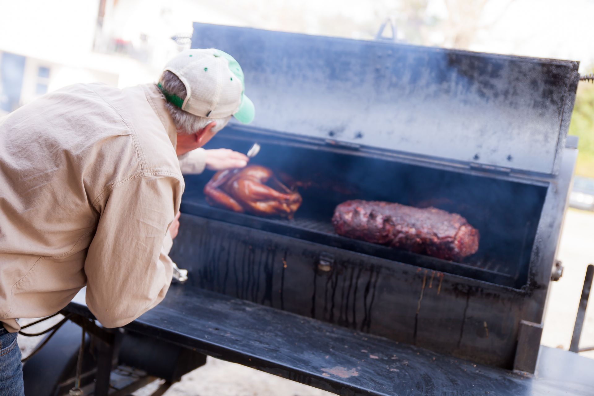 A person in a light-colored shirt and cap uses a thermometer to check the temperature of meat inside a large smoker.