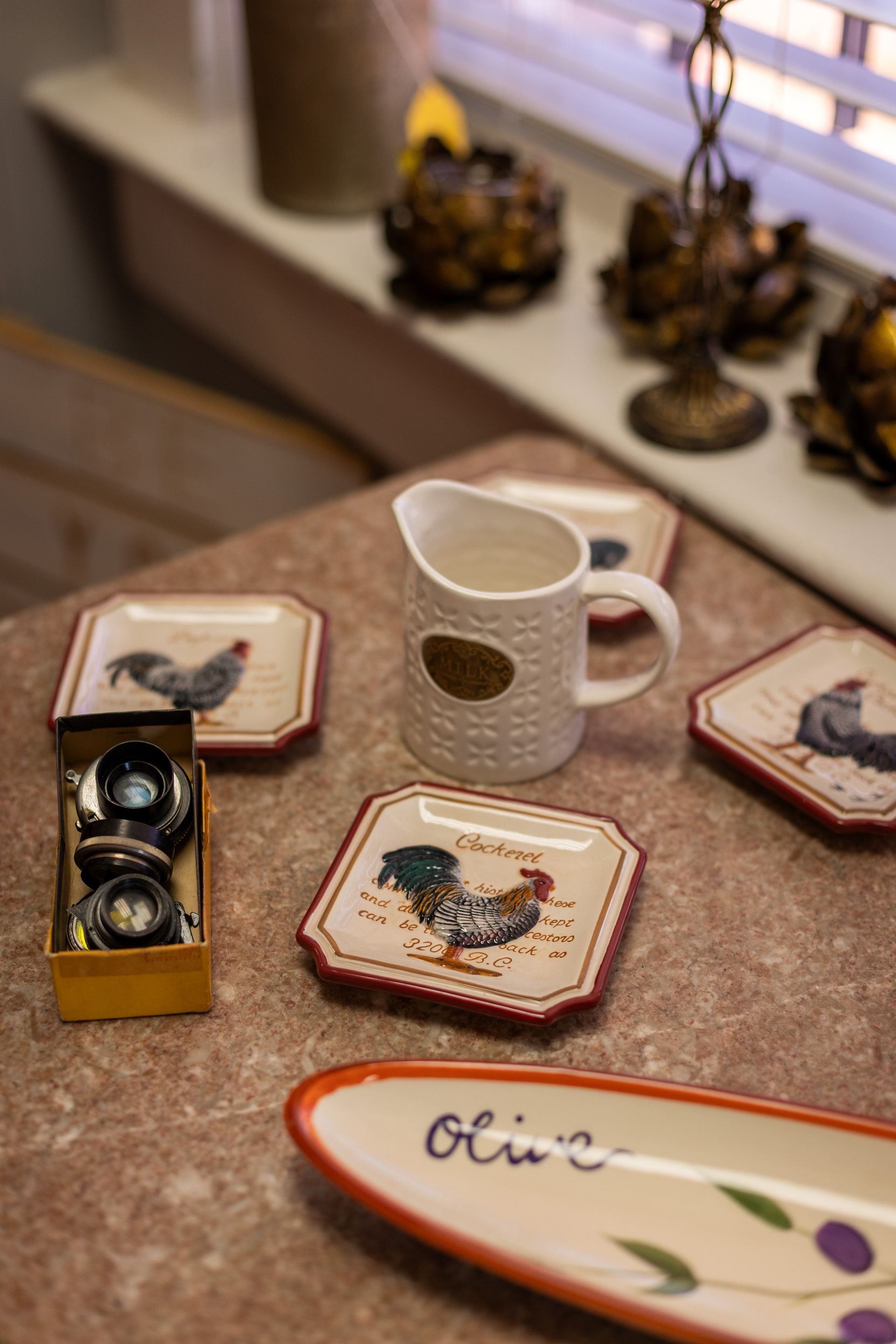 A white pitcher, rooster-themed square plates, a small box of metal hardware, and an olive-patterned tray on a table.