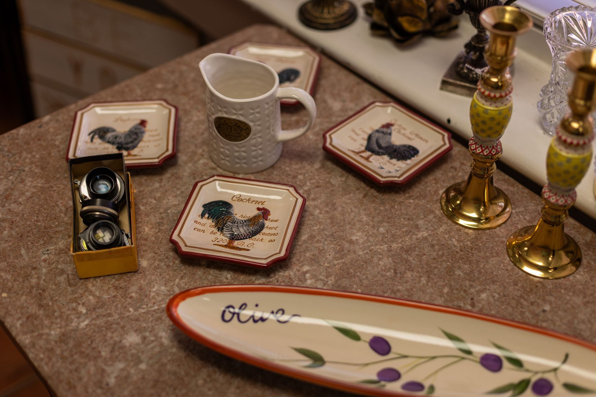 Tabletop display with rooster plates, a white pitcher, two brass candlesticks, and an olive-themed serving platter.