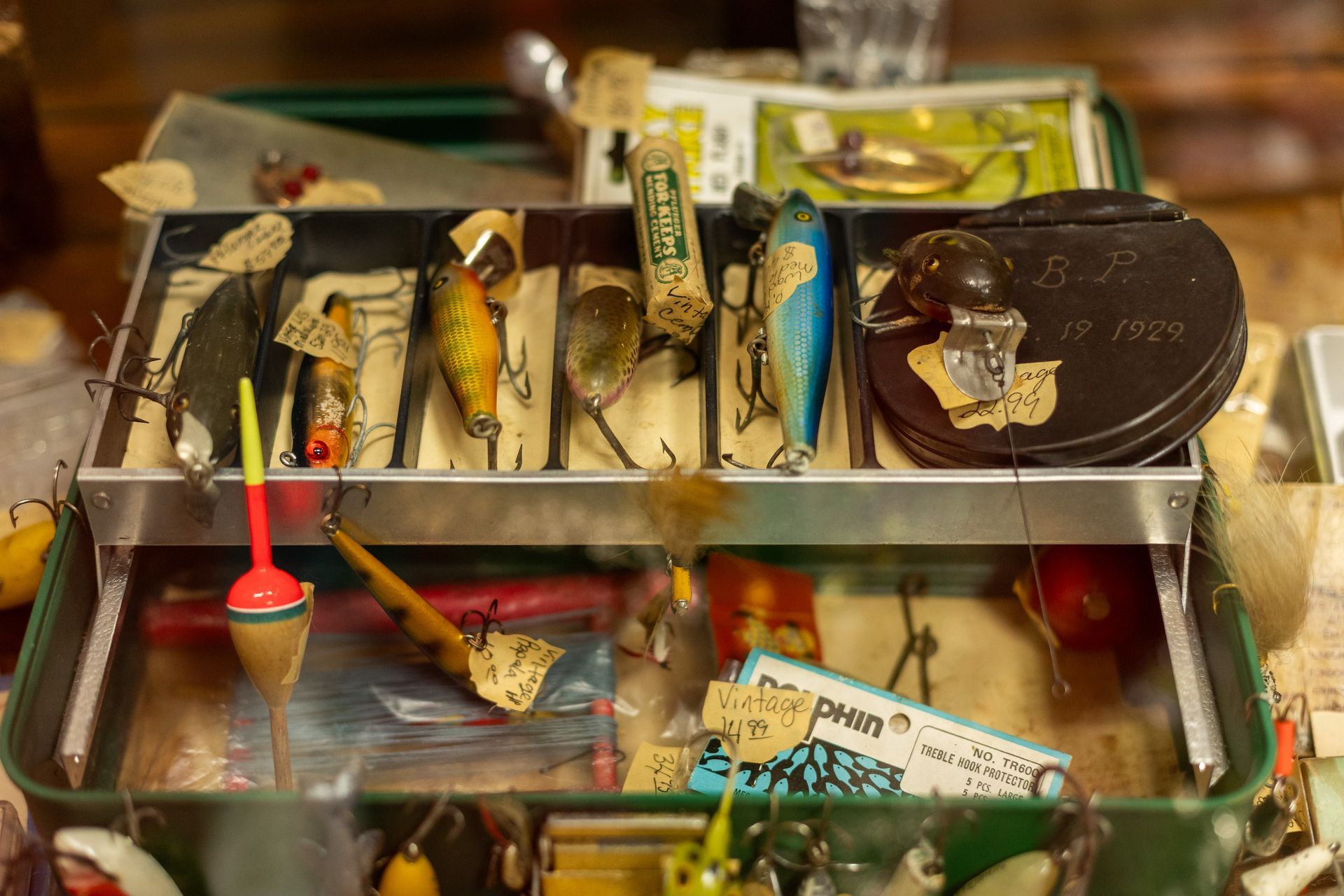 An open green metal tackle box filled with vintage fishing lures, colorful bobbers, and small packaged fishing gear.
