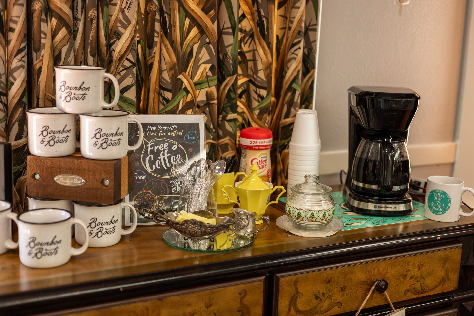 A hotel coffee station on a wooden dresser with white mugs, a coffee maker, cream, sugar, and a decorative sign.