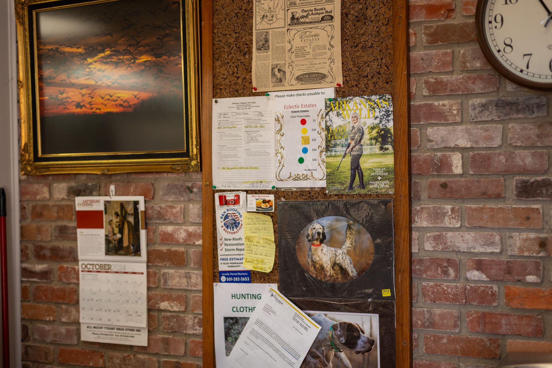 A brick wall featuring a framed sunset painting, a bulletin board with various papers and photos, and a wall clock.
