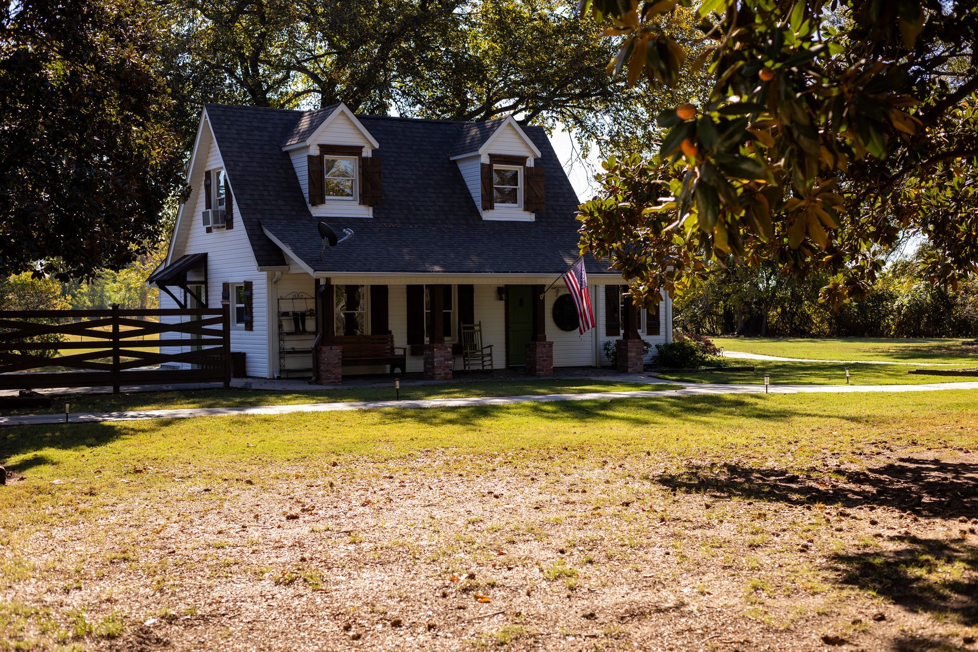 A white cottage with dormer windows, a front porch, and an American flag, set on a grassy lawn with surrounding trees.