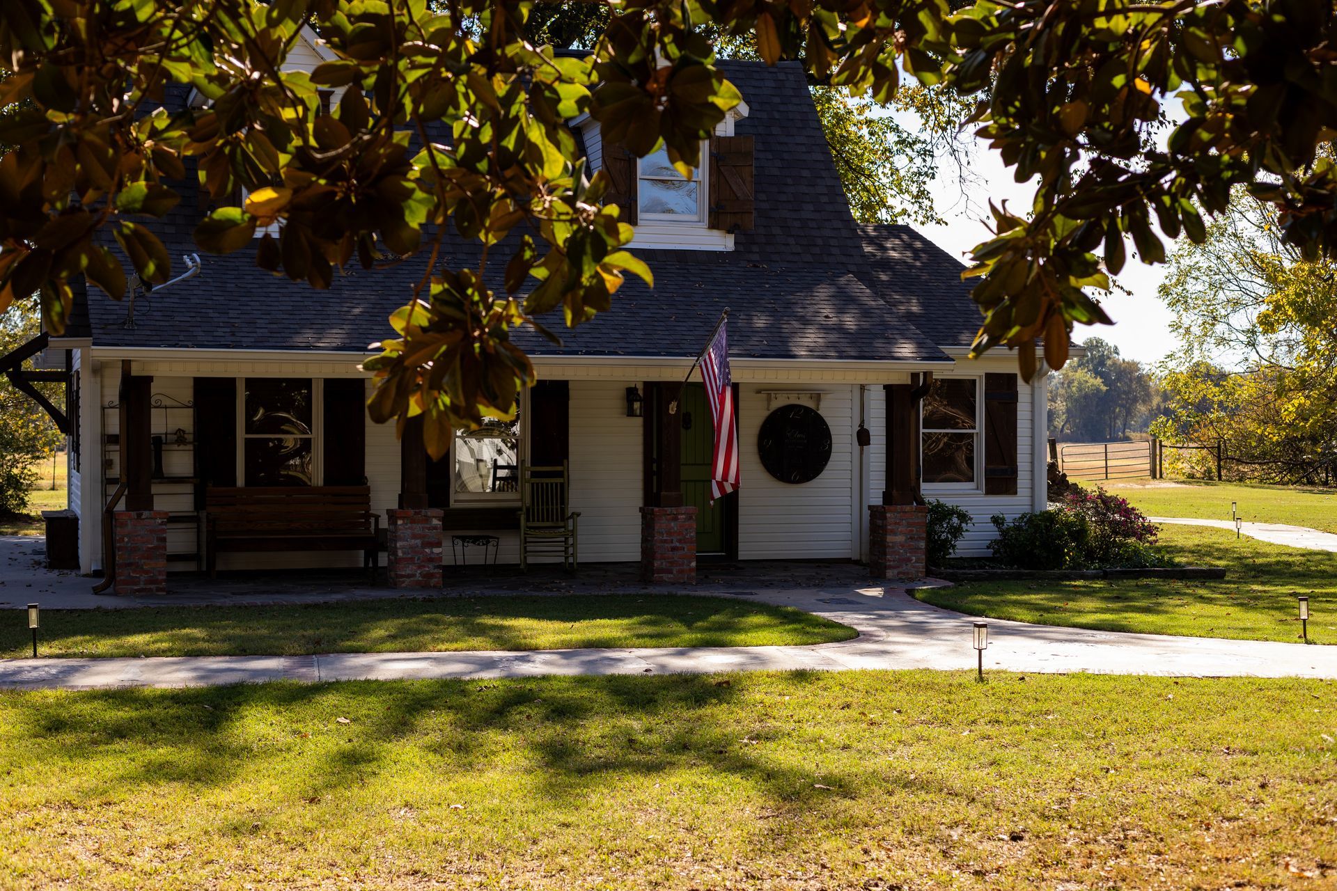 A white, single-story house with dark shutters and a front porch, viewed from a grassy yard under tree branches.