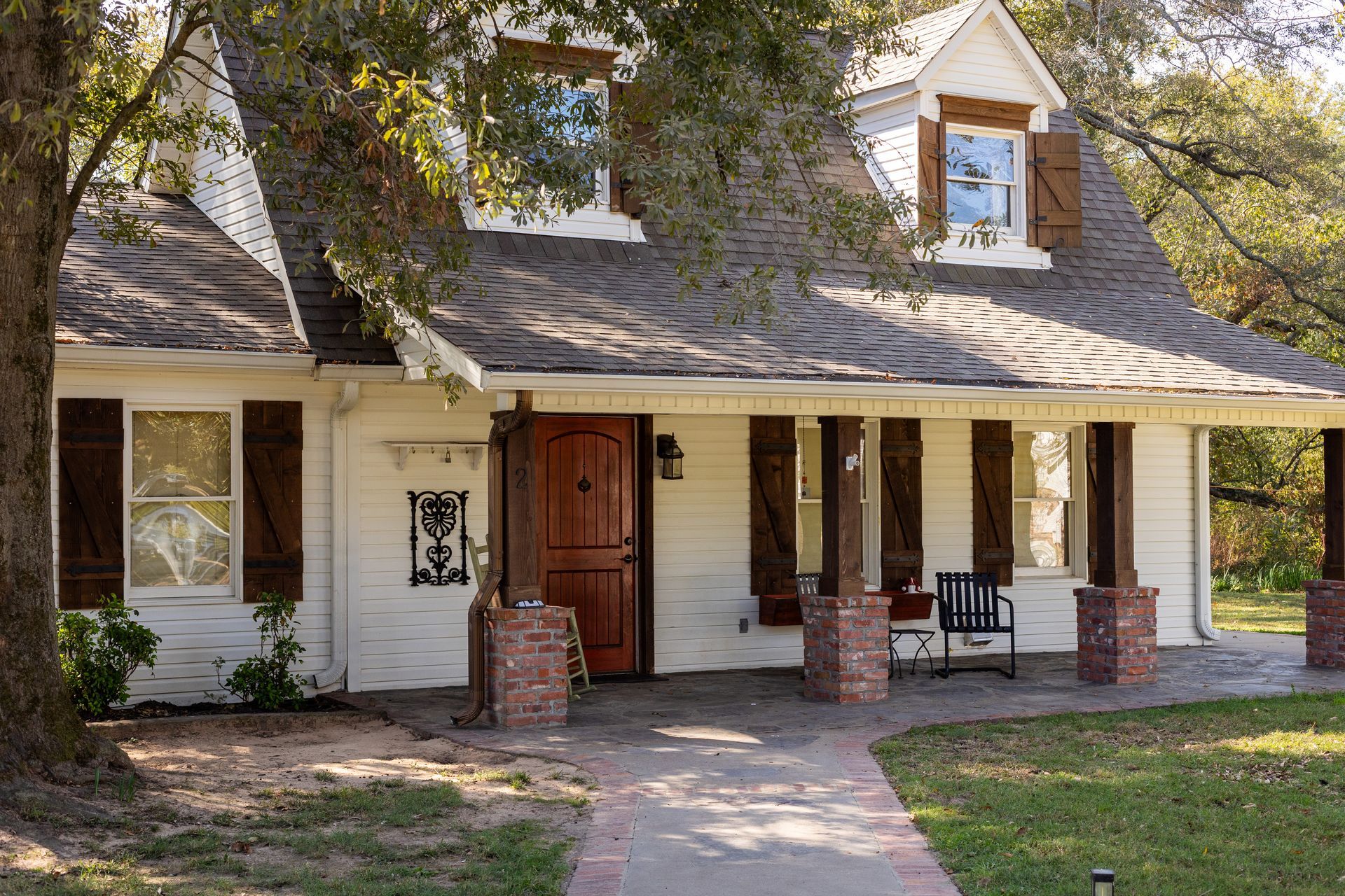 A white, one-story house with a dormer, dark brown shutters, a wooden front door, and a brick-pillared covered porch.