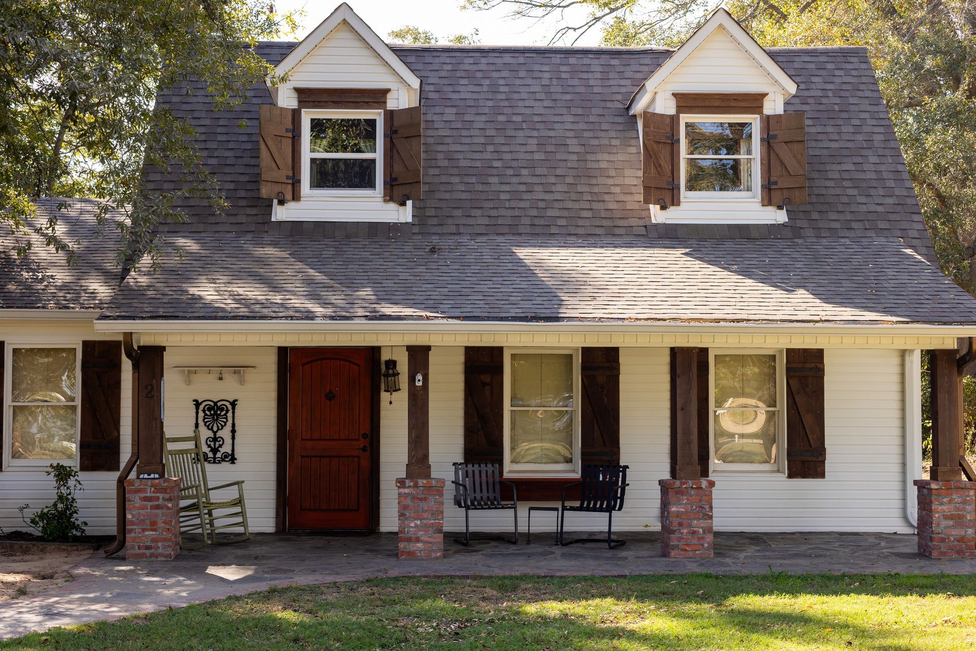 A white Cape Cod style house with a dark roof, wooden shutters, brick-based columns, and a front porch.
