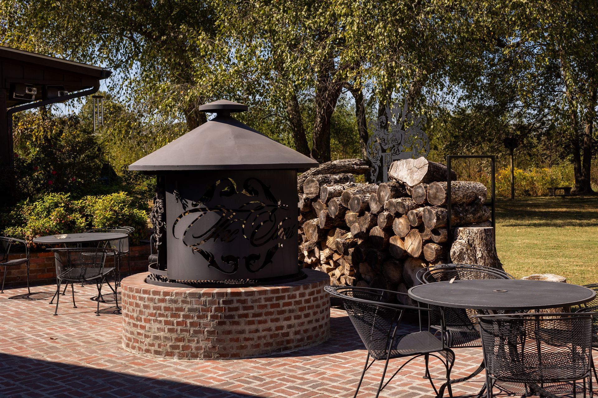 A black metal fire pit sits on a circular brick patio, surrounded by outdoor furniture and a stack of firewood nearby.
