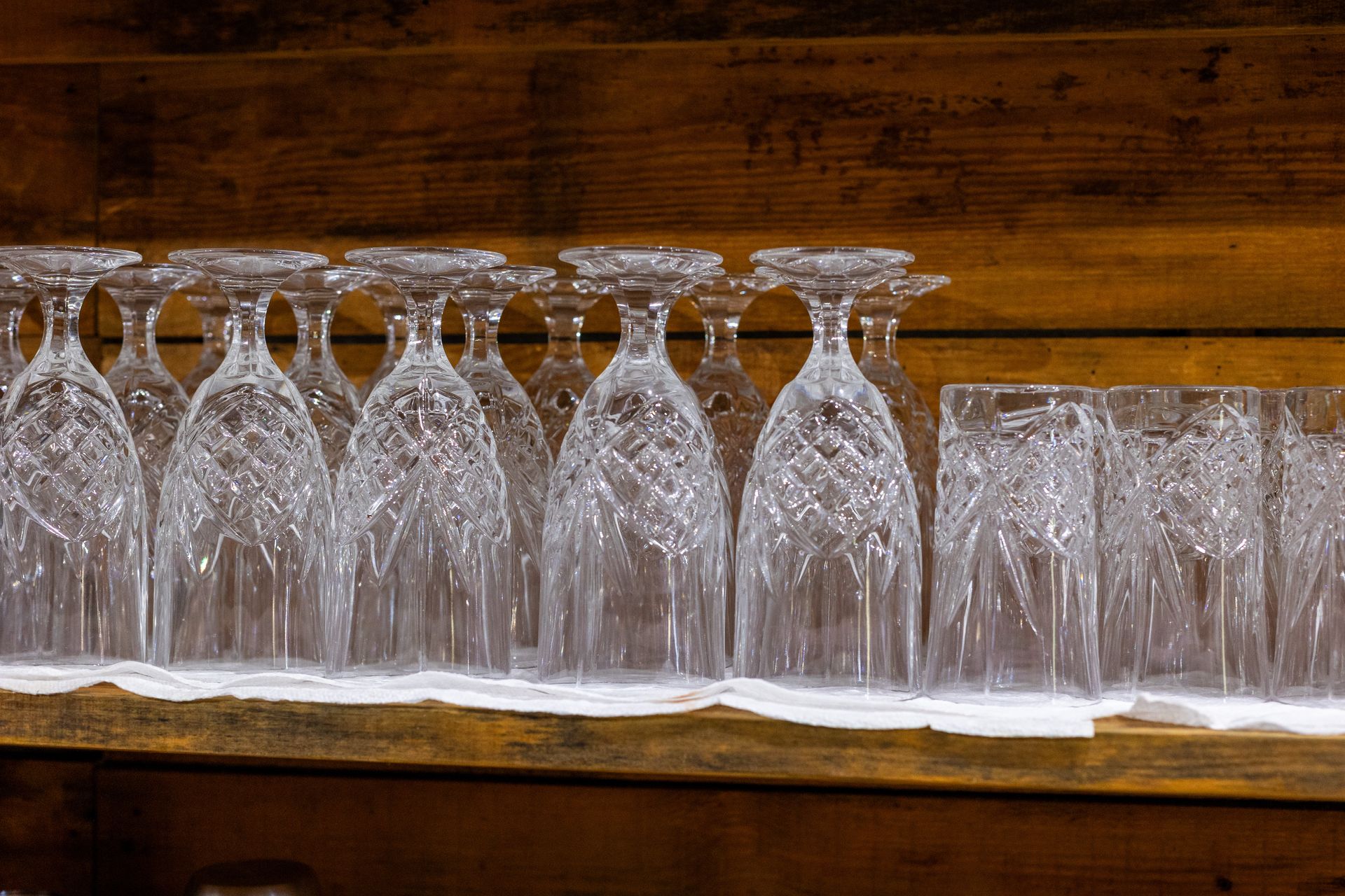 Clear patterned glasses are lined up on a wooden shelf against a wooden wall.