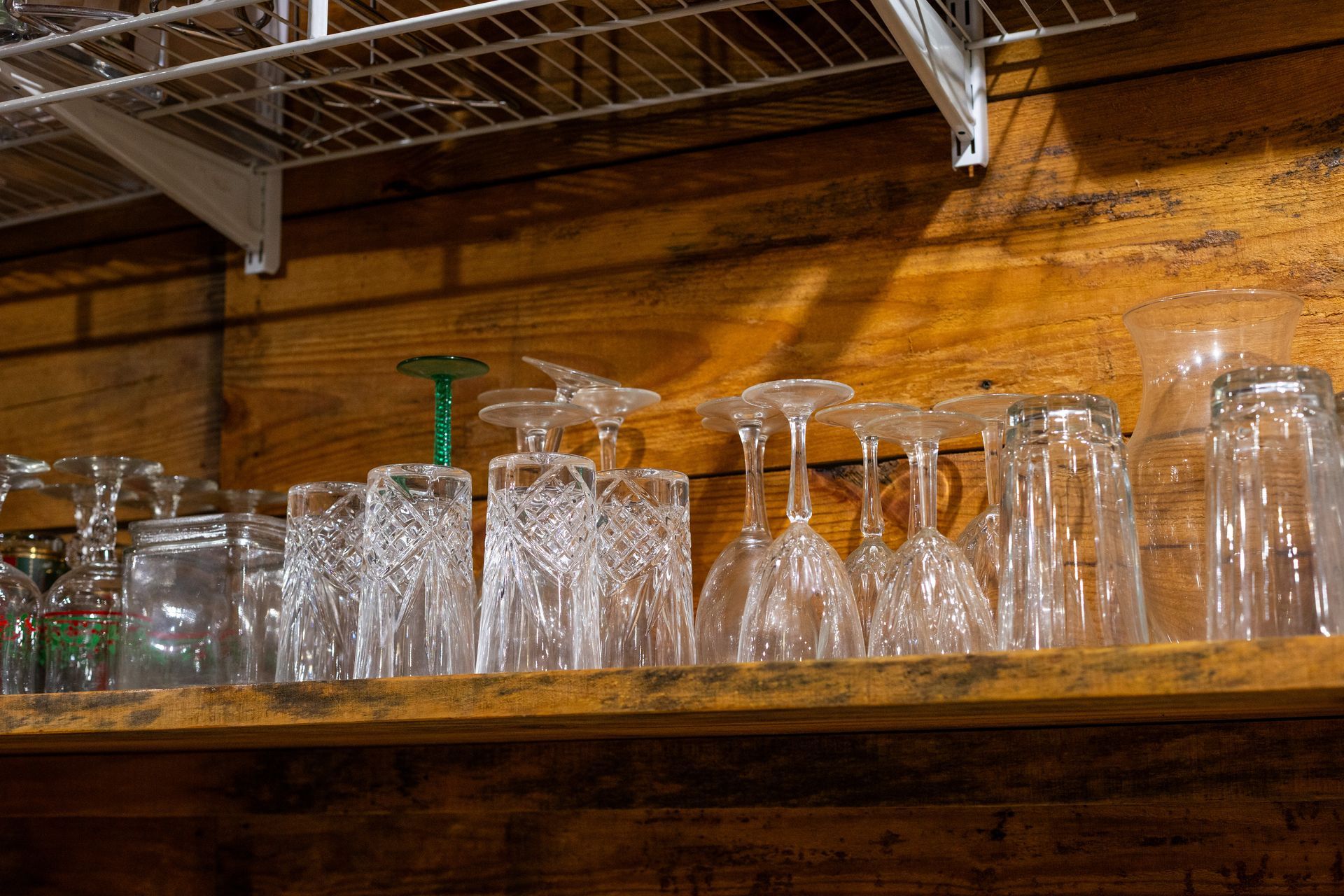 Various glass tumblers and wine glasses stored upside down on a wooden shelf against a wooden wall.