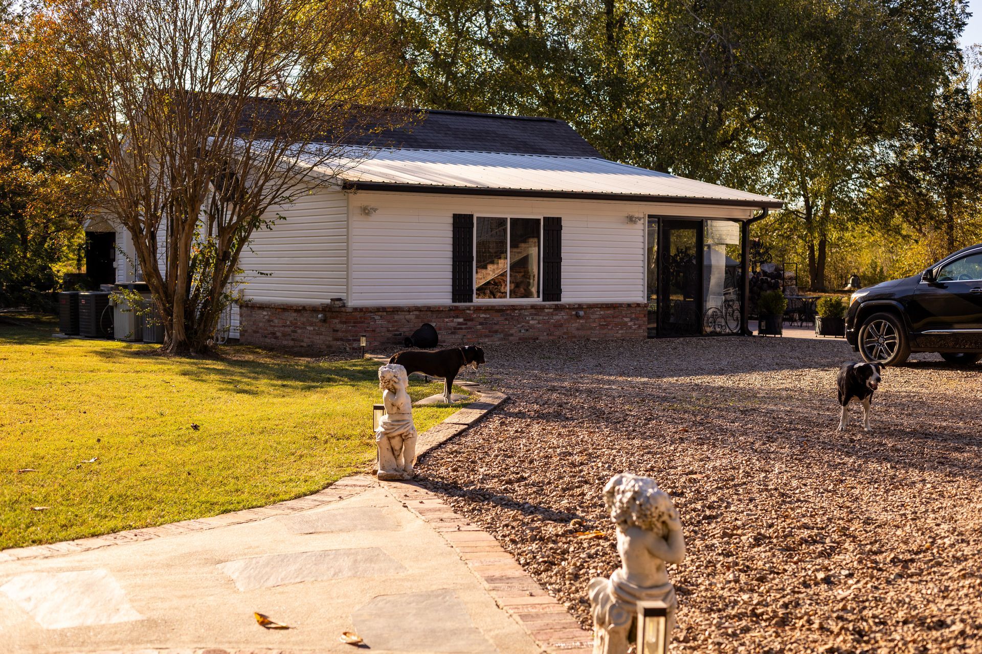 A white home with a brown roof and brick foundation sits on a gravel driveway with stone statues and dogs on the lawn.