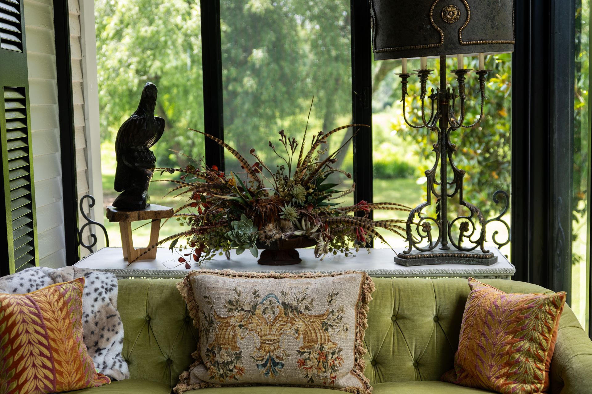A green tufted sofa sits in front of a windowed porch, featuring decorative throw pillows, a flower arrangement, and lamp.