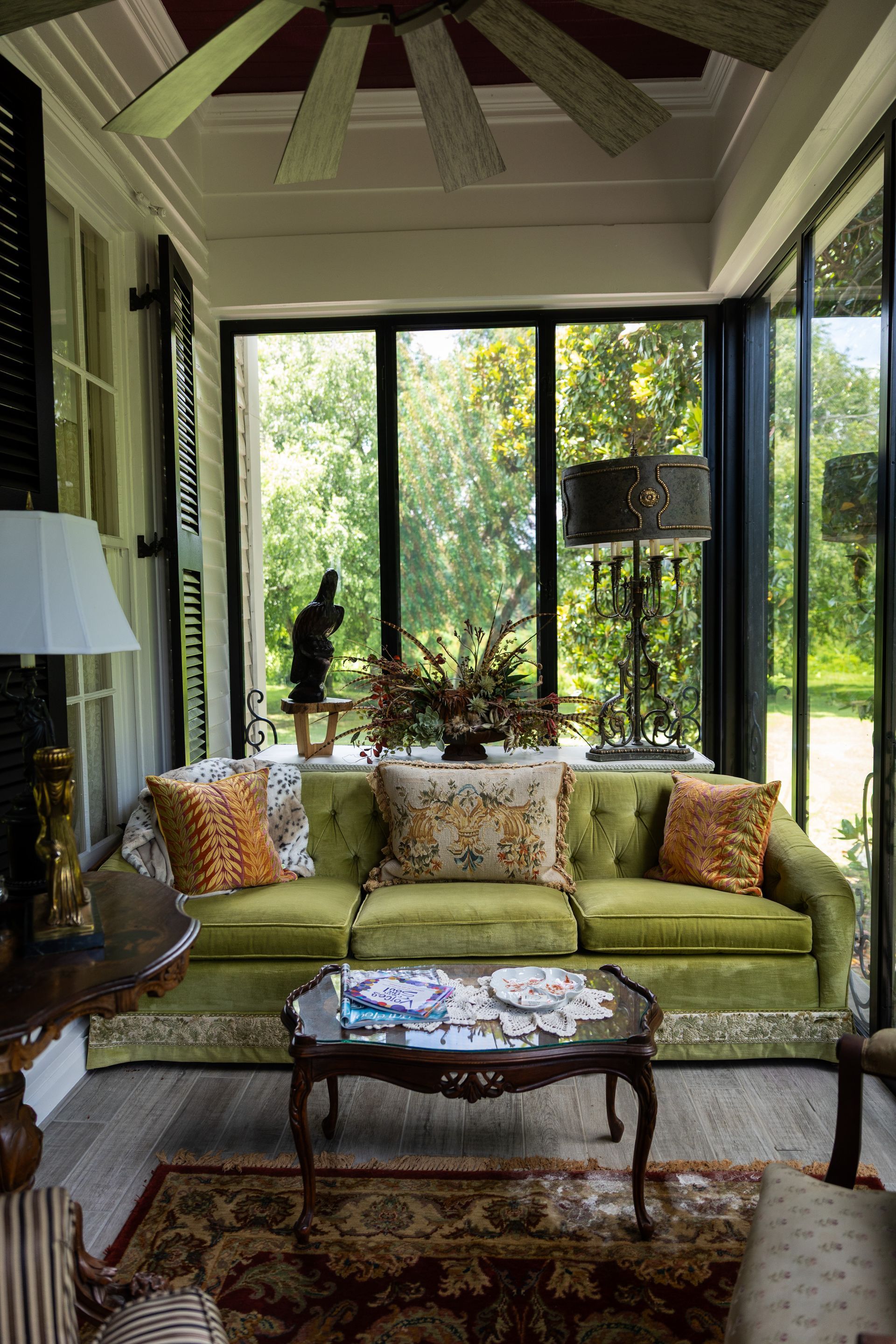 A sunroom with a green tufted sofa, floral and orange pillows, a glass coffee table, and large windows with garden views.