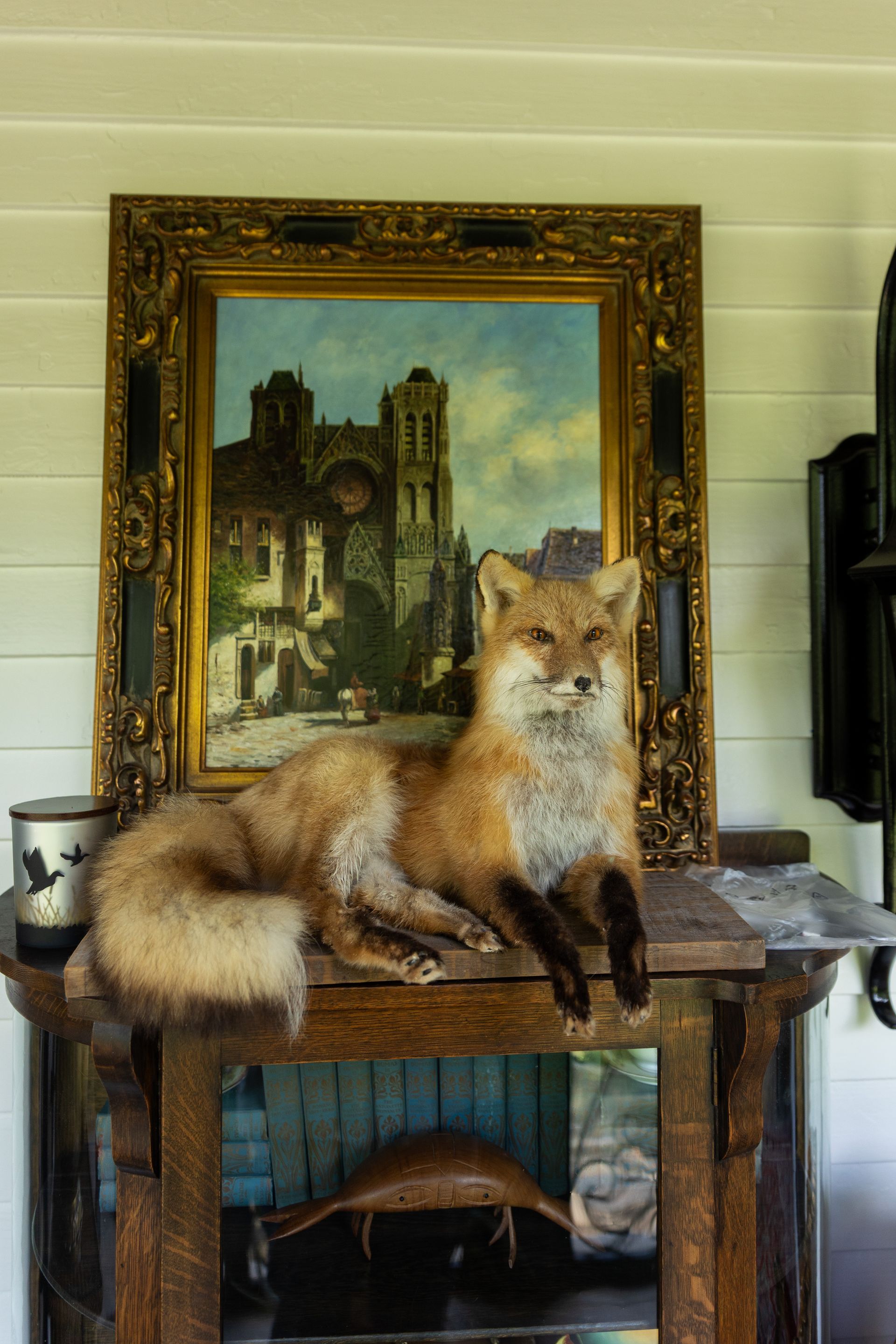 A taxidermy red fox resting on a wooden stand in front of a framed painting of a cathedral.