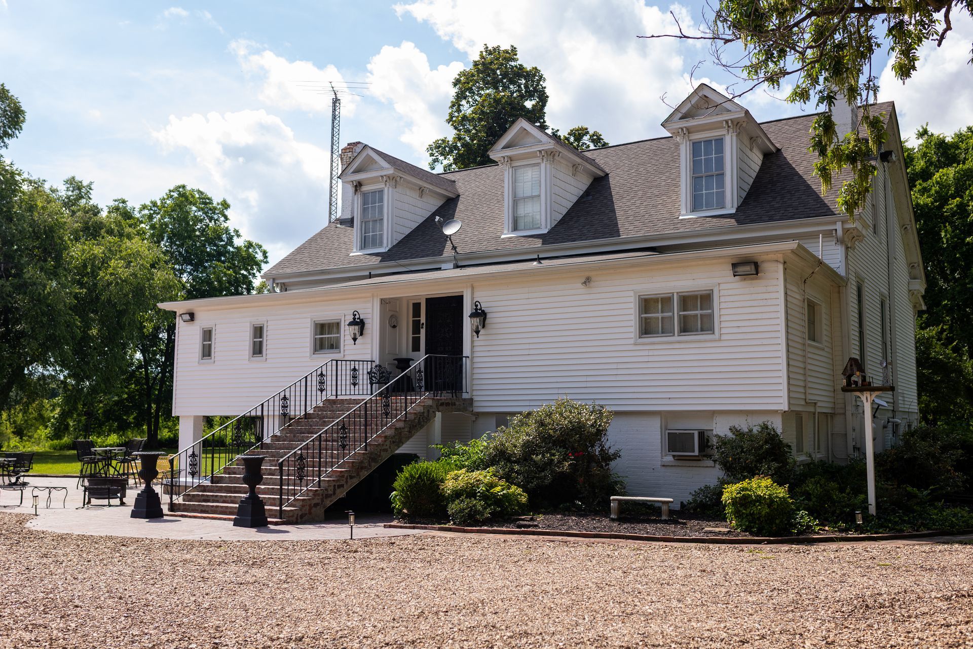 A two-story white house with dormer windows, a front stone staircase, and a gravel yard under a partly cloudy sky.