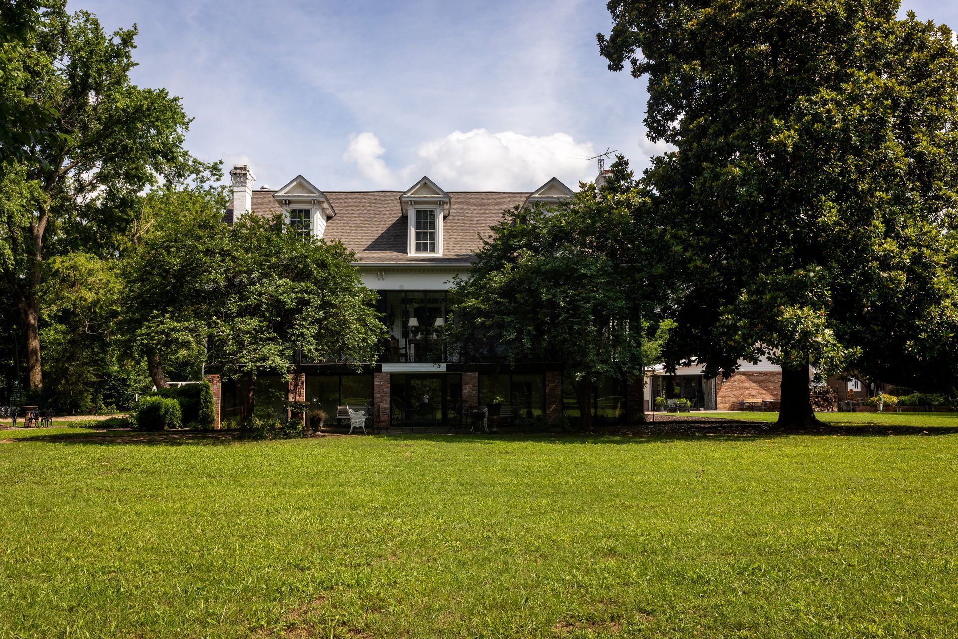 A large, two-story house with a dormer roof and glass ground level, surrounded by trees and a wide, green lawn.