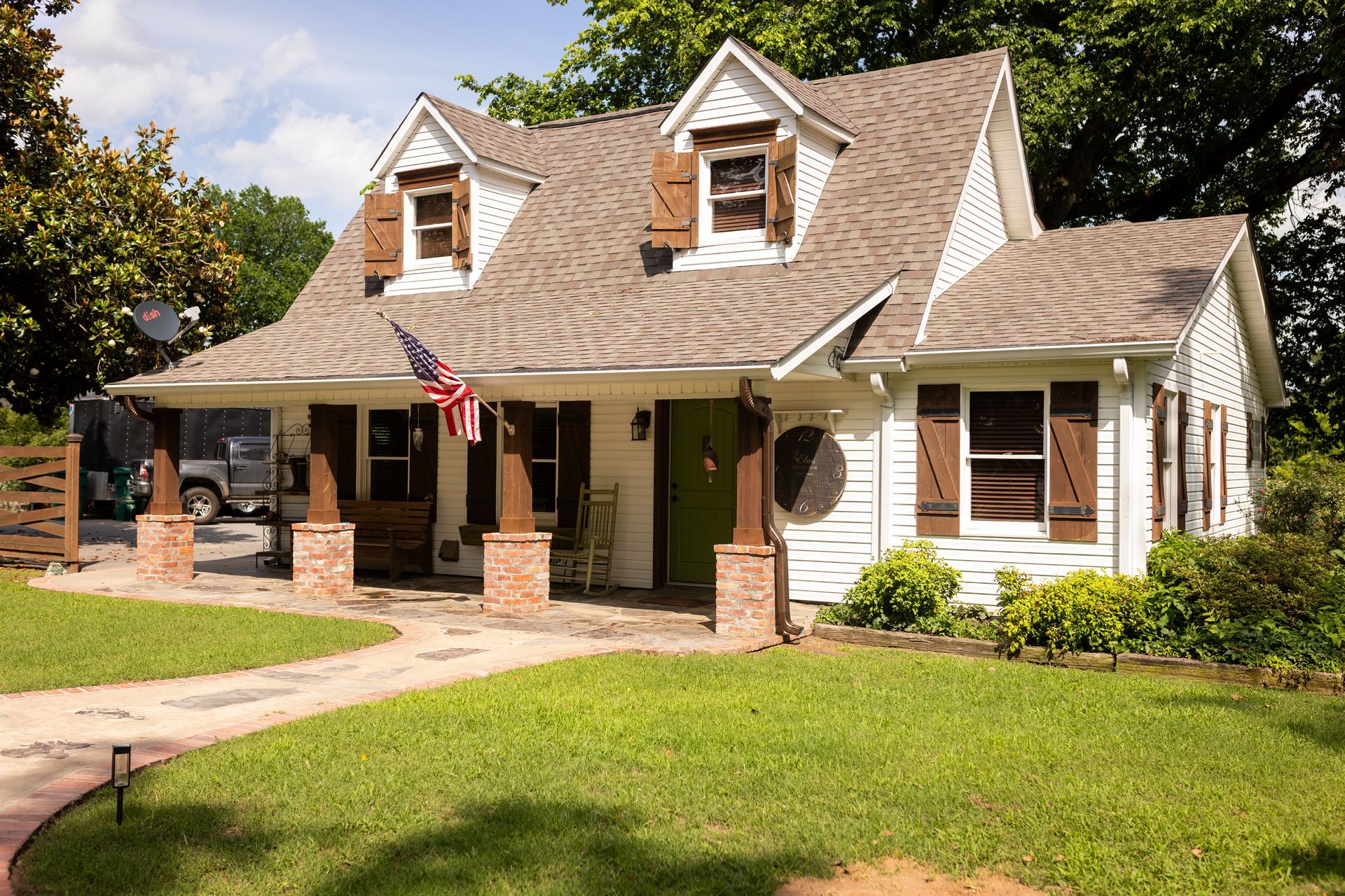 A white cottage-style house with brown shutters, a green front door, and a porch, set in a yard with a stone walkway.