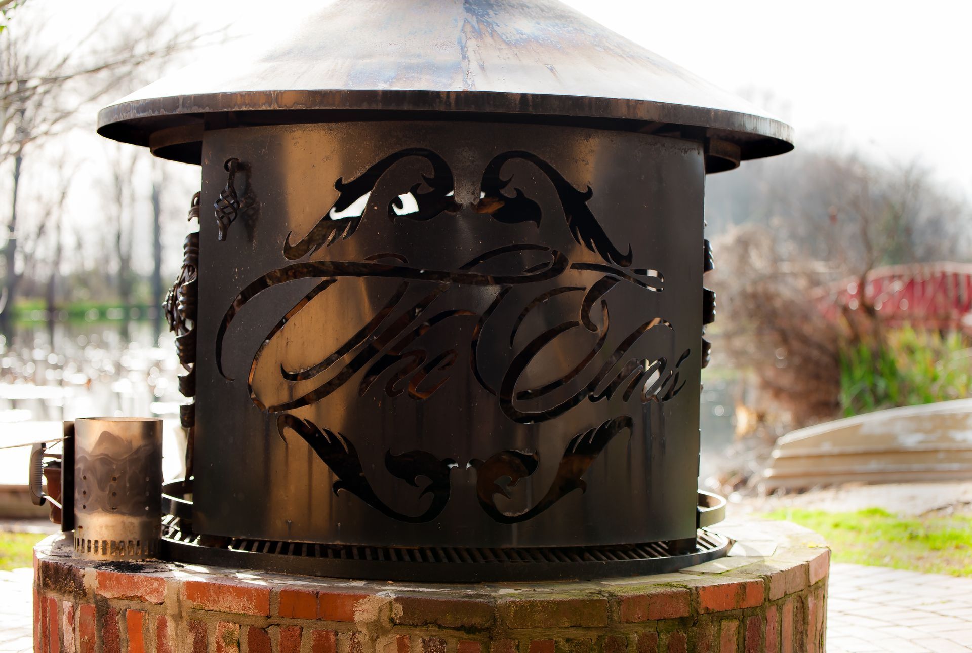 A black metal fire pit with custom cutouts sits on a brick base outdoors near a pond.
