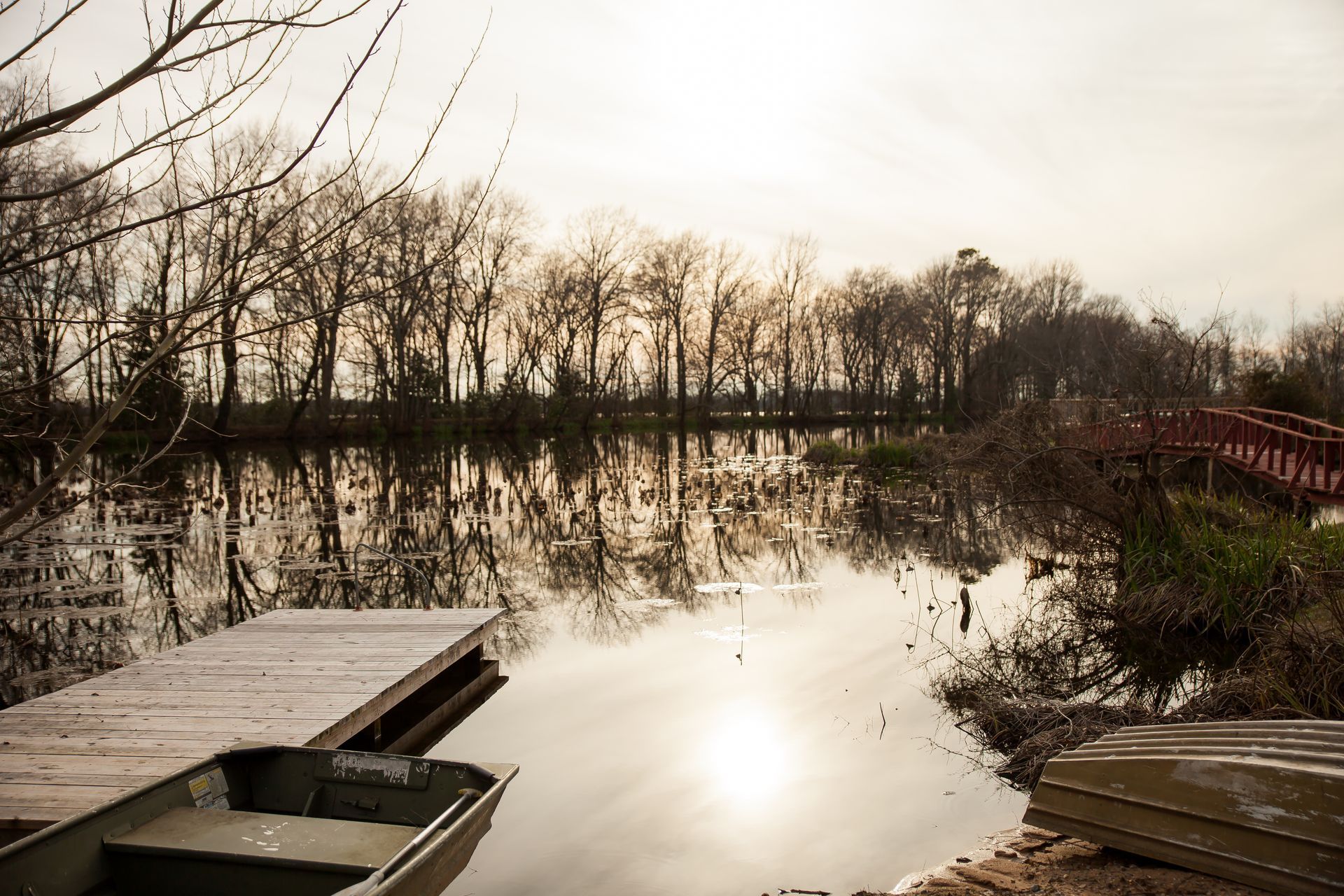 A wooden dock extends into a calm, reflective river at sunset, with bare trees along the bank and a small boat nearby.