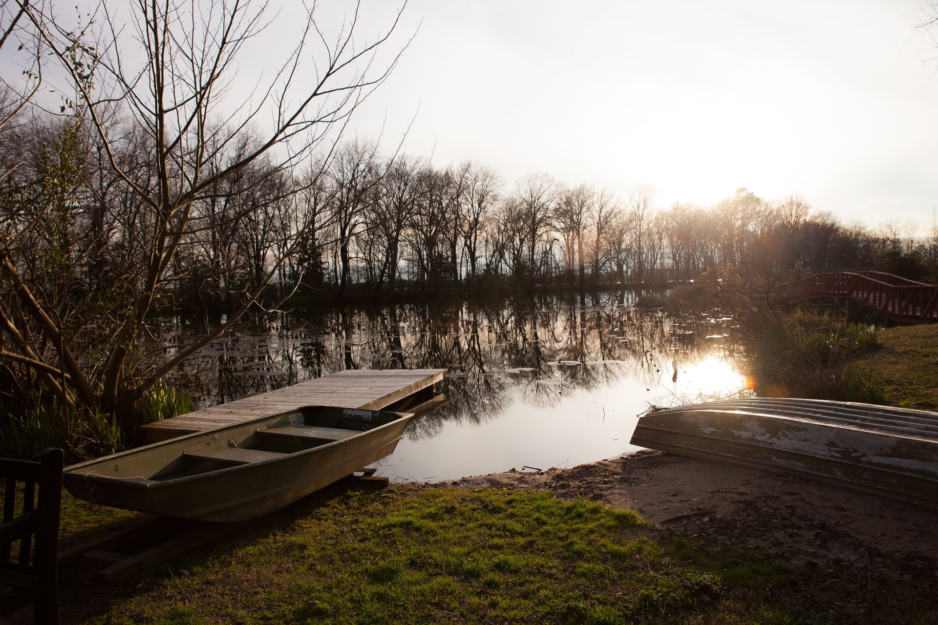 A boat rests beside a wooden dock on a calm lake at sunset, with a line of bare trees reflected in the water.