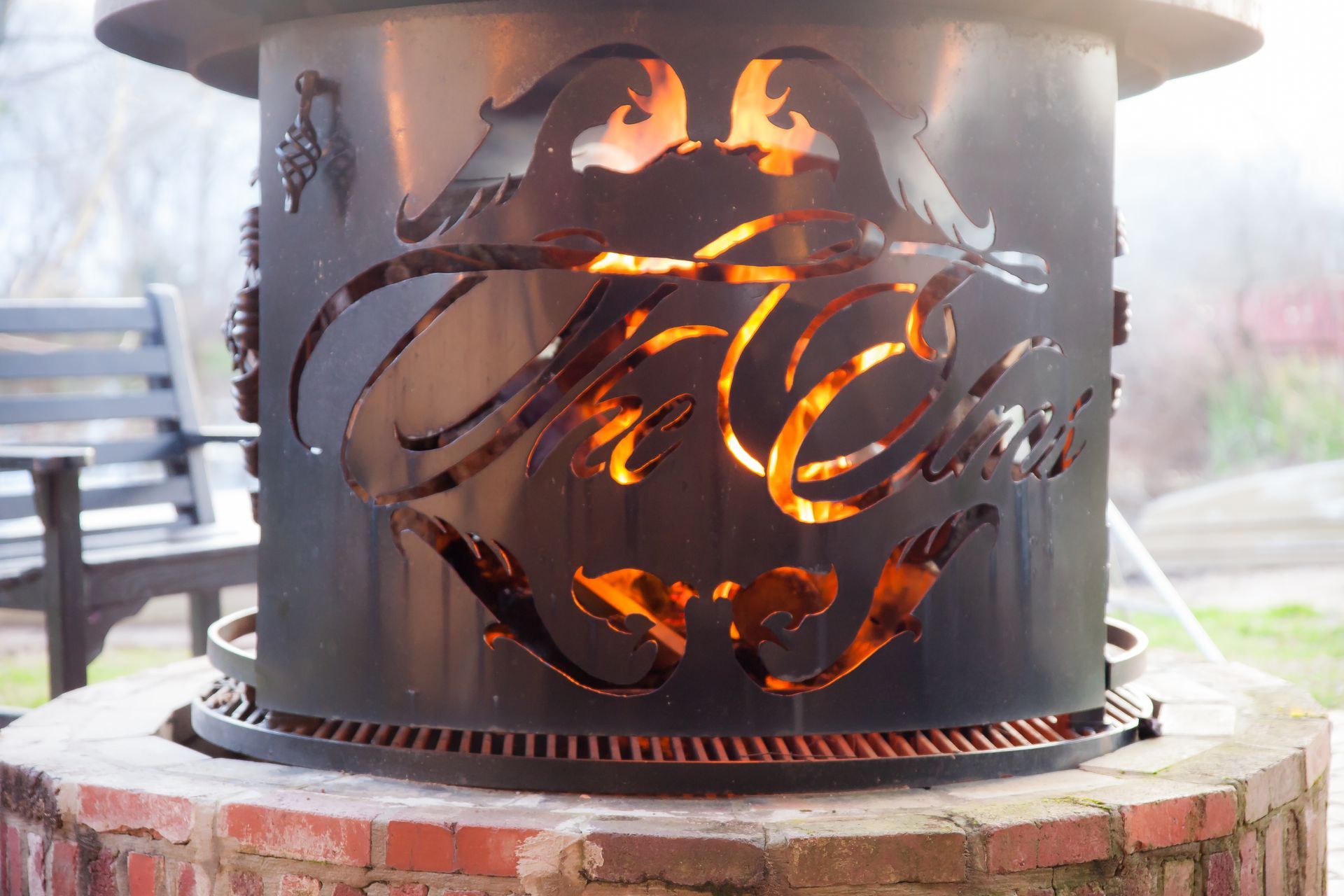 A metal fire pit ring with decorative cutouts glowing from the fire within, set on a brick base outdoors.