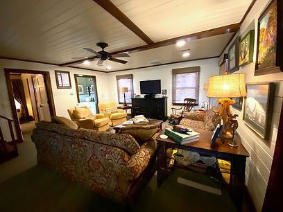 A cozy living room with a patterned sofa, two armchairs, a television, a floor lamp, and wall art on a wood-paneled wall.