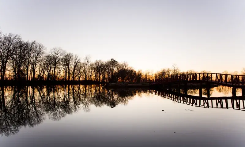 A wooden footbridge stretches across a calm lake at sunset, reflecting the bare trees and warm sky on the water's surface.