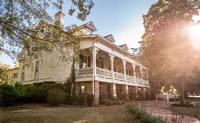 A classic two-story white house with a wraparound porch, surrounded by lush trees under a bright, sun-drenched sky.