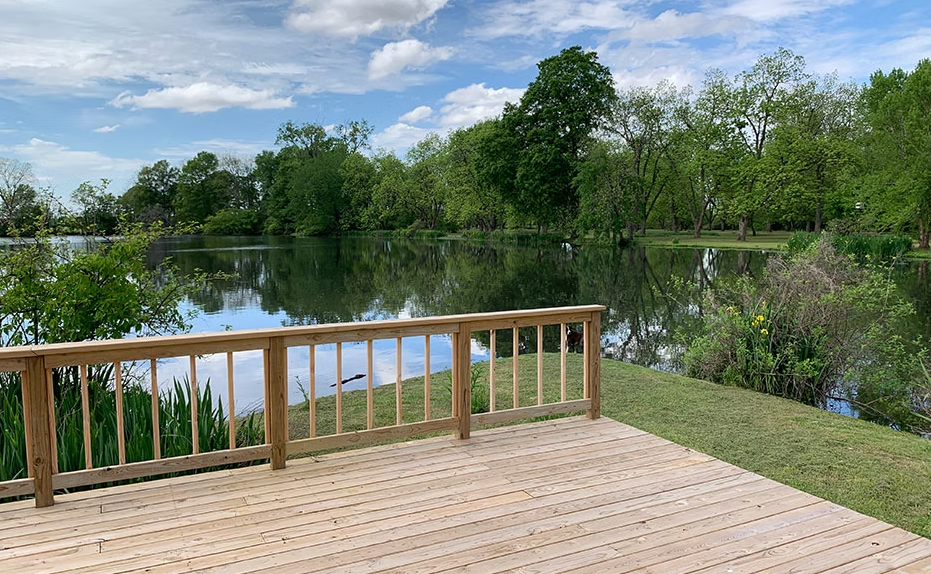 A new light-wood deck with simple railings overlooking a calm pond surrounded by lush green trees under a cloudy blue sky.