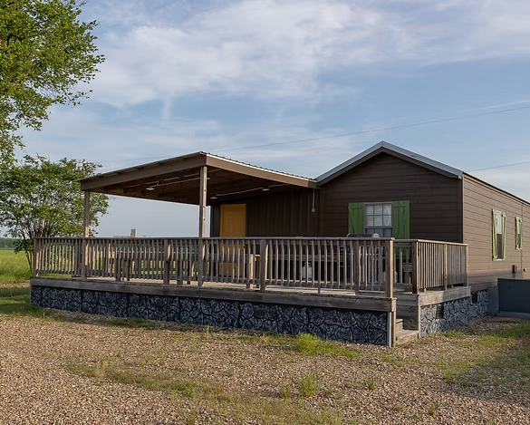 A brown modular home with a large wooden deck, a covered patio area, and a dark stone-patterned skirting on a gravel lot.