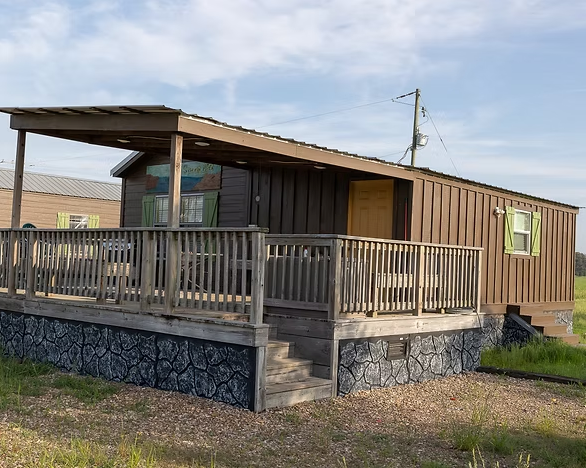 A brown, single-wide manufactured home with a large wooden porch and a stone-patterned foundation under a clear sky.