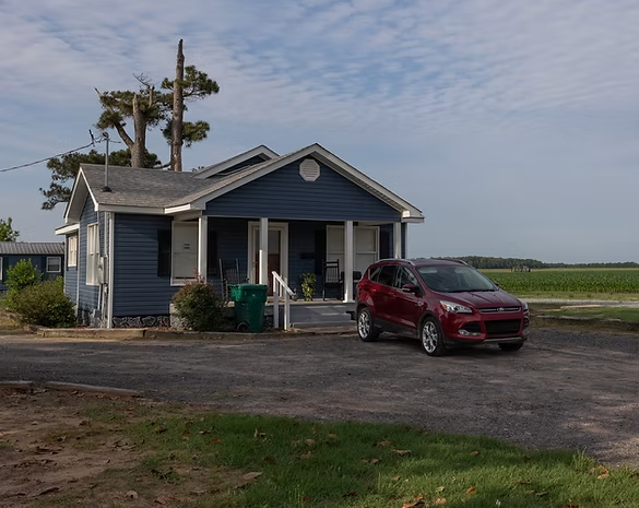 A small blue house with a porch and a red car parked in the driveway in front of an open, grassy field.
