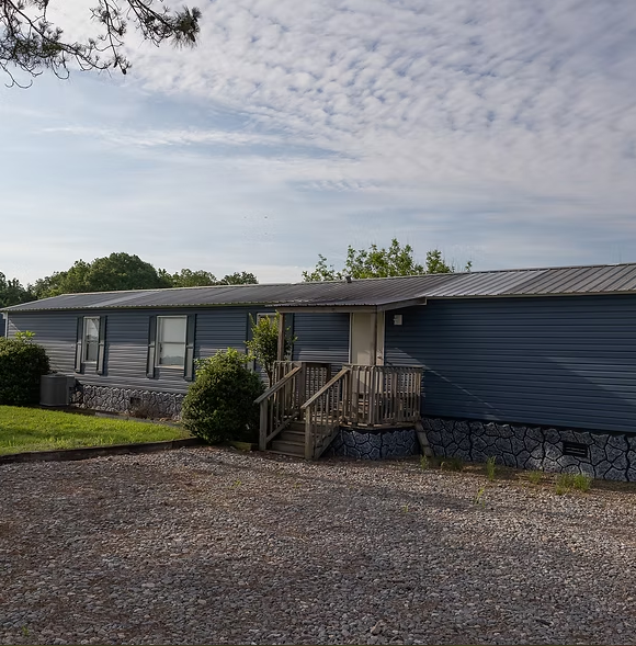 A blue manufactured home with a metal roof and a small wooden porch sits on a gravel lot under a cloudy sky.