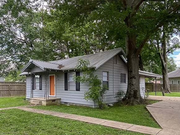 A one-story gray house with a metal roof, orange front door, and a concrete path, set beside a large tree on a grassy lot.