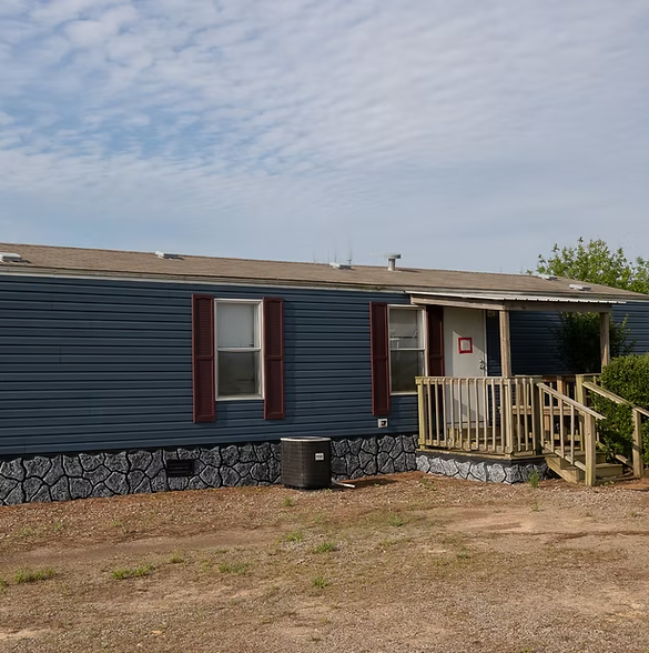 A blue mobile home with brown shutters, a wooden porch, and faux-stone skirting set against a partly cloudy sky.