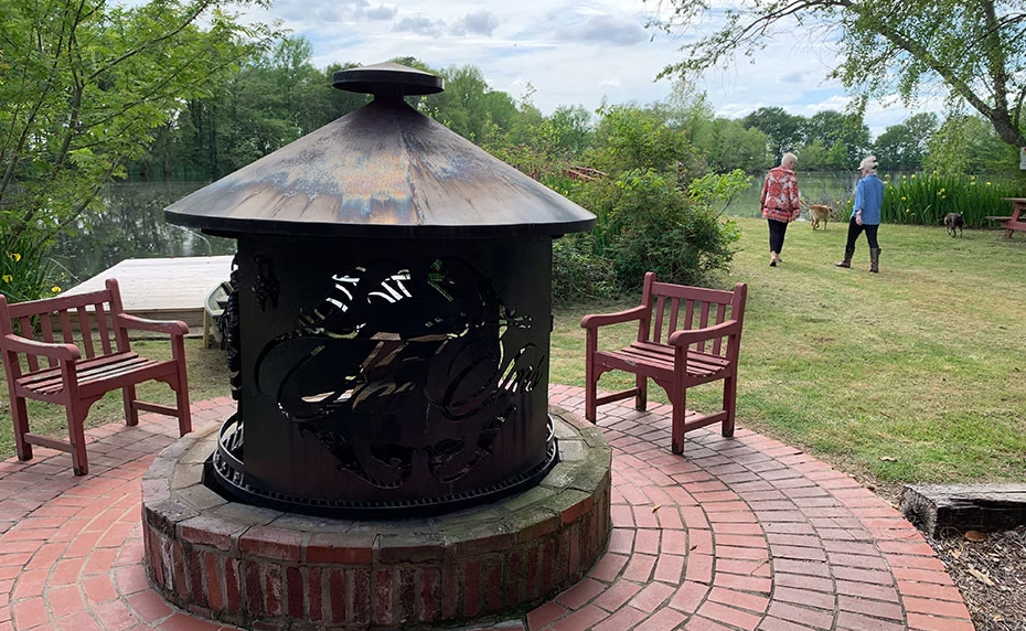 A metal fire pit sits on a circular brick patio with two wooden chairs, overlooking a river with two people in the yard.