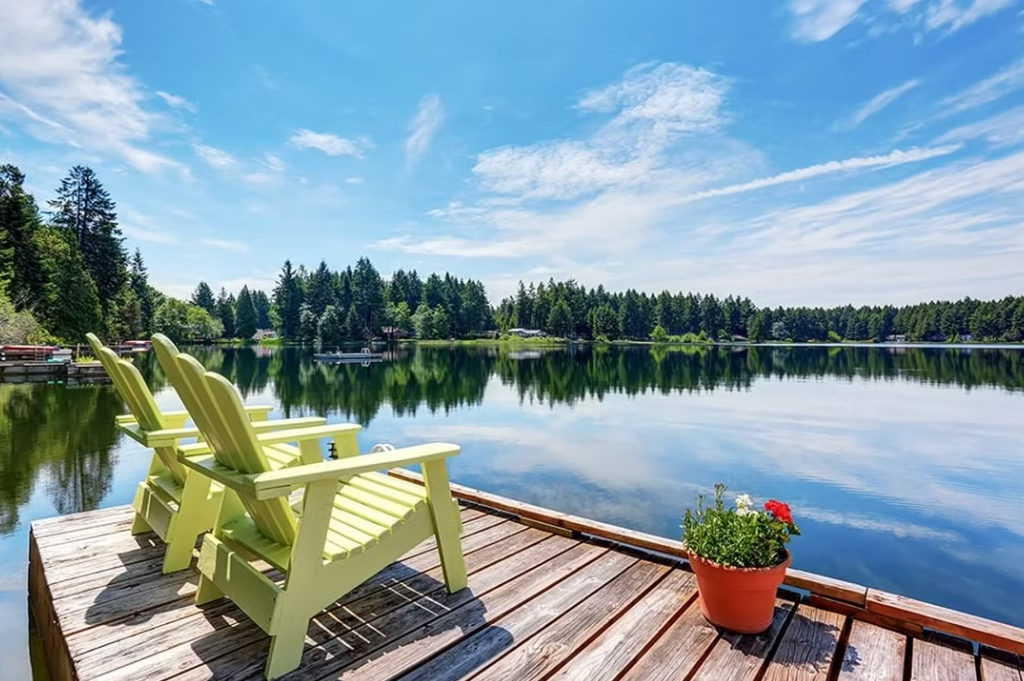 Two light green Adirondack chairs on a wooden dock overlooking a calm lake surrounded by trees under a blue sky.