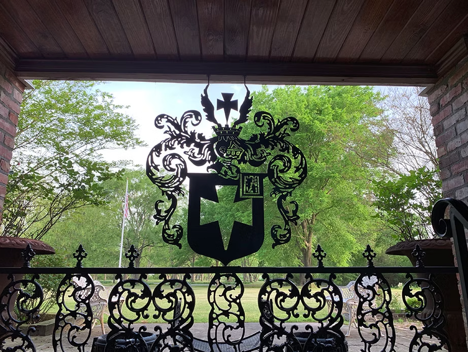 Black metal gate featuring a central coat of arms with a cross, set against a blurred background of trees and a lawn.